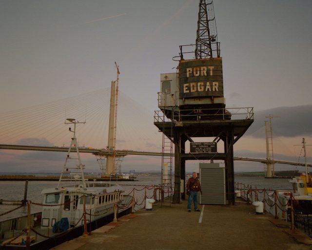 A man stands in front of a large boat crane at Port Edgar harbour during blue hour